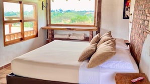Une chambre avec un grand lit, une tête de lit en bois, une fenêtre donnant sur un paysage verdoyant et un mur en briques.