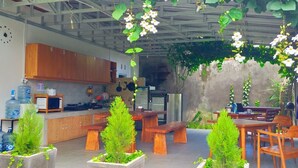 Un área de patio al aire libre con mesas y bancos de madera, rodeada de plantas en macetas y flores colgantes.
