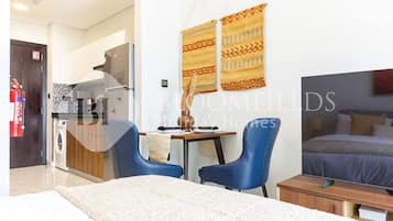 A modern hotel room with a bed, a desk, two chairs, a kitchen area, and a fire extinguisher.