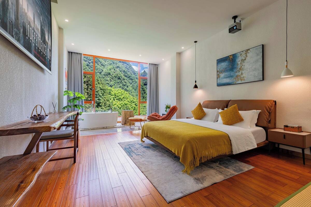 Quarto moderno com uma cama grande, uma escrivaninha de madeira e vista para vegetação exuberante.