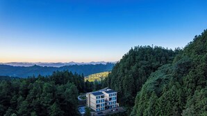 A modern building with a flat roof and multiple windows, surrounded by dense forest and mountains in the background. - Huagu Jianzhu Guesthouse Zhangjiajie (Zhangjiajie)