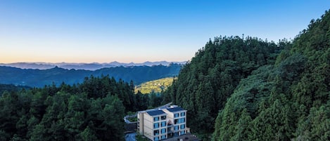 A modern building with a flat roof and multiple windows, surrounded by dense forest and mountains in the background.