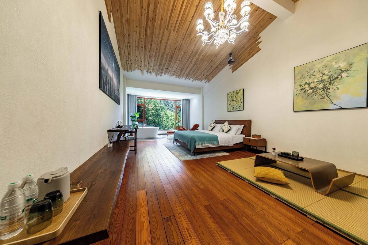 Quarto de hotel moderno com teto de madeira, cama com manta verde, sofá e vista para o exterior.
