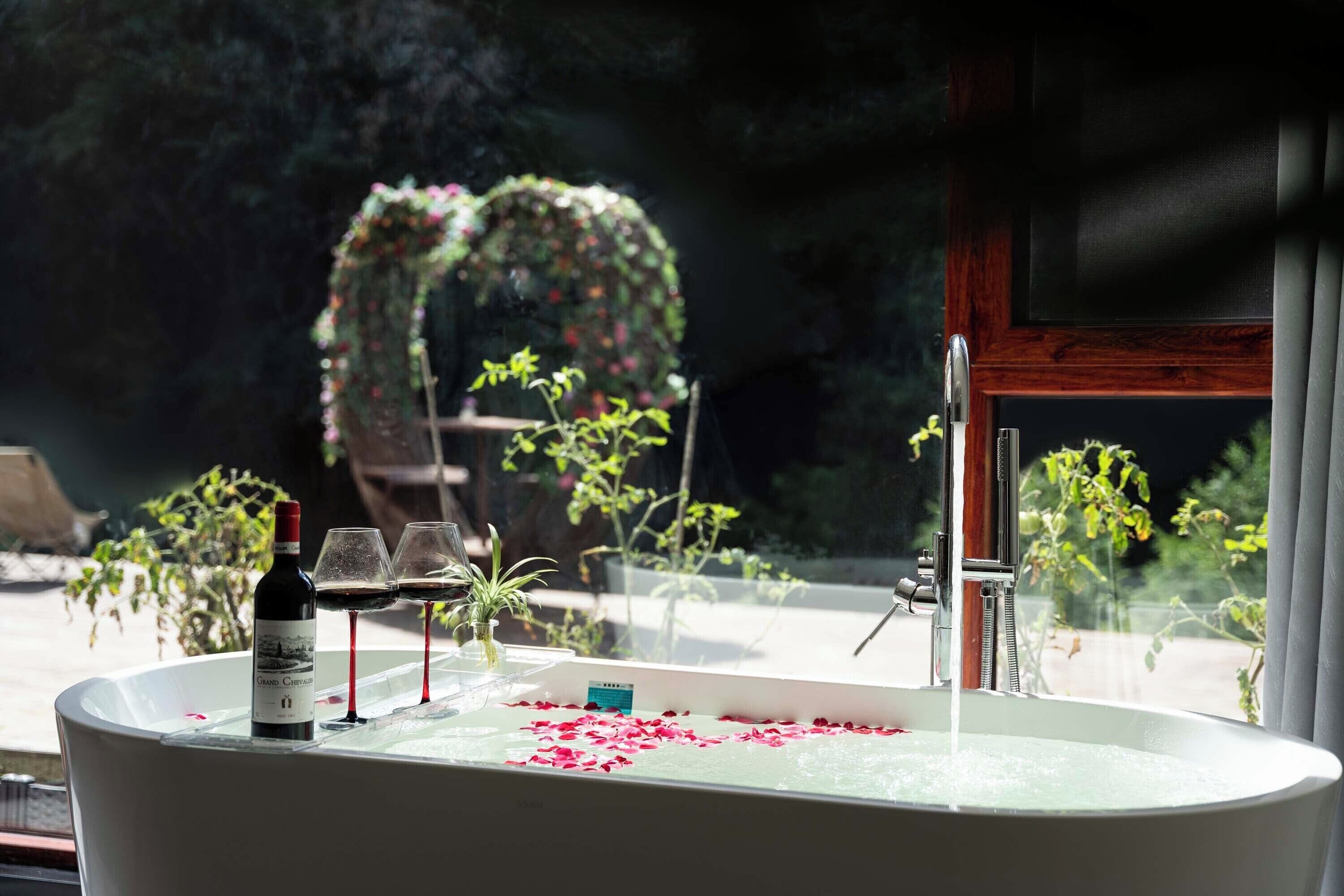 Una bañera con pétalos de rosa, una botella de vino y copas sobre una mesa.