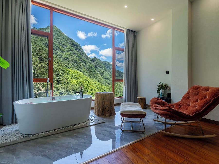 Ein modernes Badezimmer mit einer freistehenden Badewanne, einem roten Schaukelstuhl und einem großen Fenster mit Blick auf eine gebirgige Landschaft.