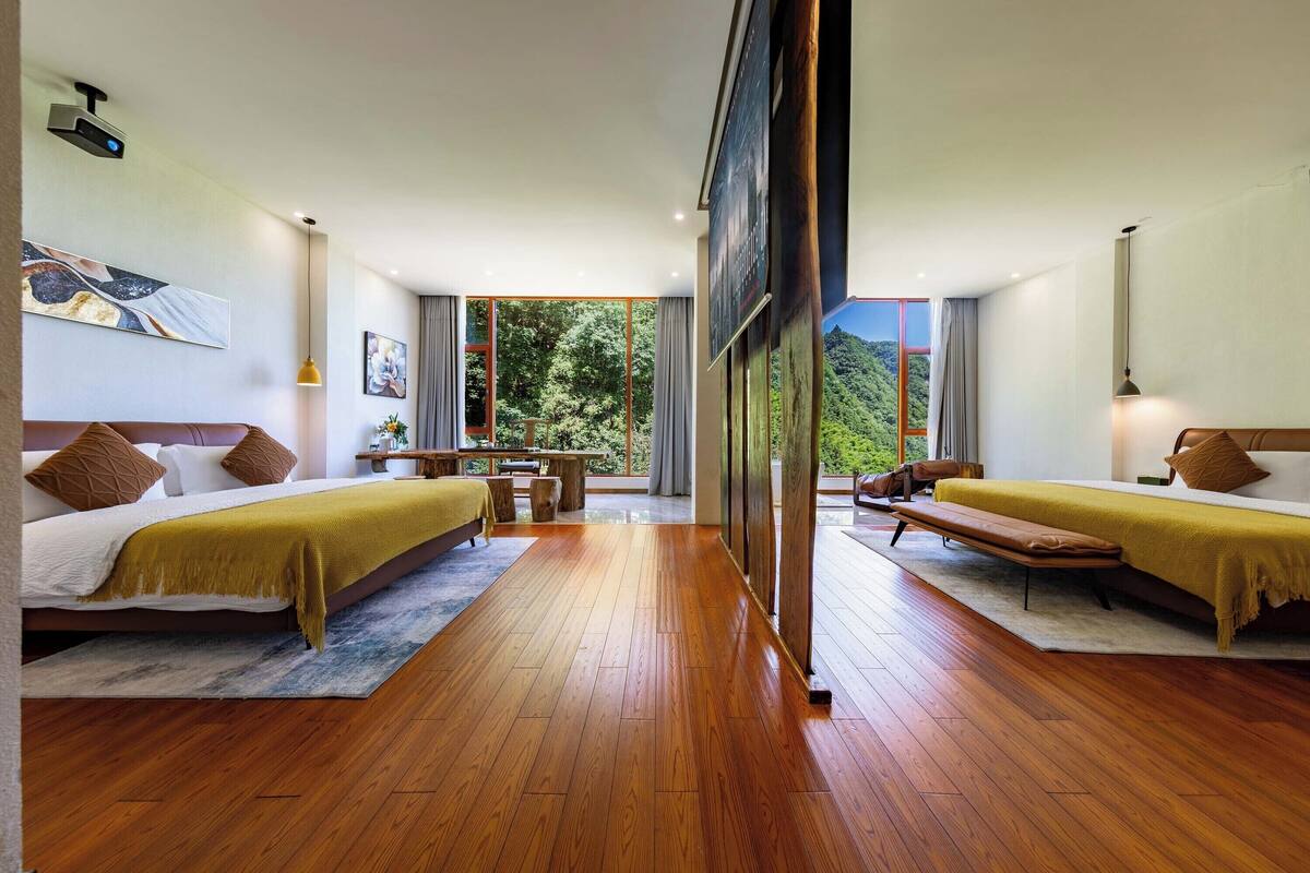 Quarto de hotel moderno com duas camas, piso de madeira, uma grande janela com vista para as montanhas e área de refeições com mesa e cadeiras.