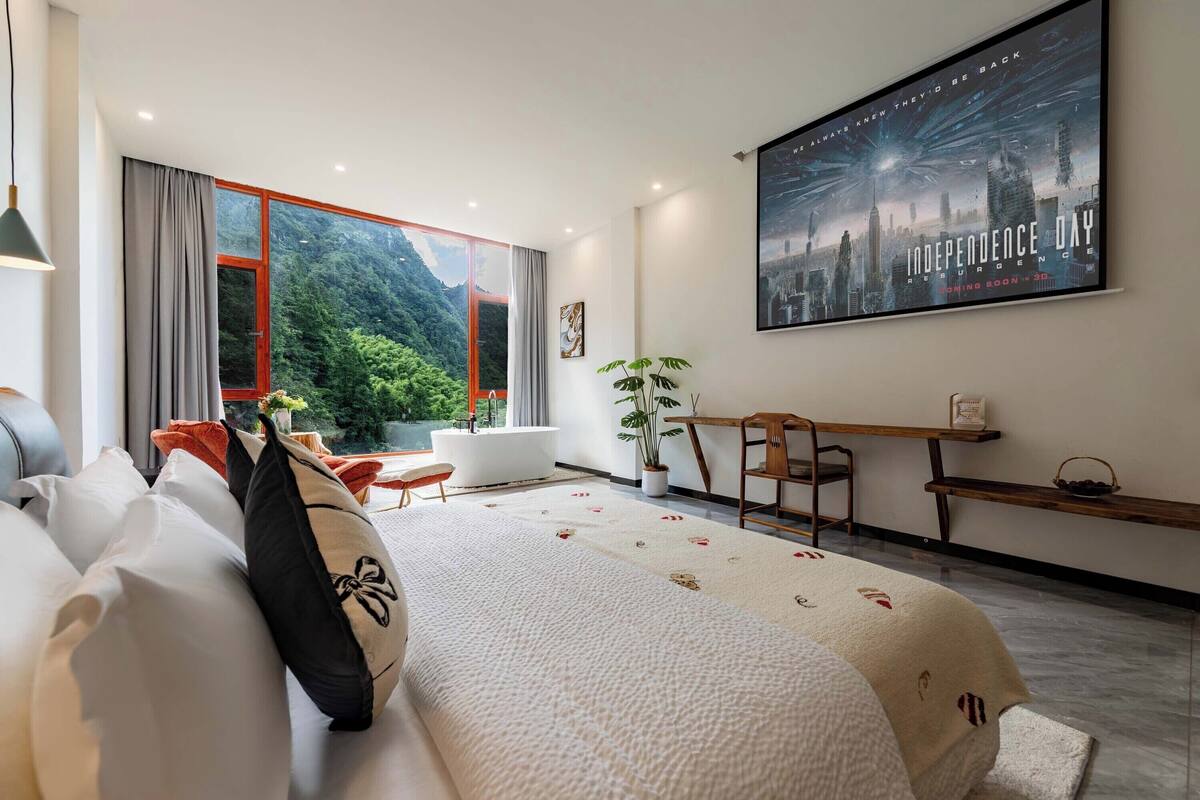 Quarto de hotel moderno com uma cama grande, uma escrivaninha, uma cadeira e uma janela ampla com vista panorâmica das montanhas.