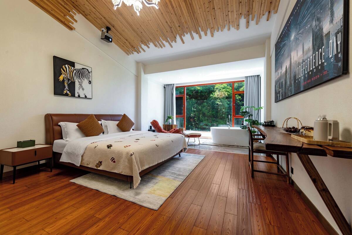 Quarto moderno com piso de madeira, uma cama com colcha estampada, uma mesinha de cabeceira, uma escrivaninha com cadeira e uma grande janela com vista para vegetação.