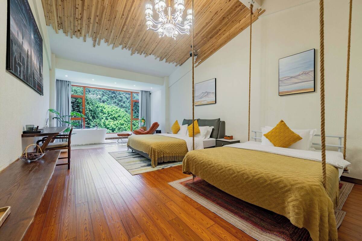 Quarto moderno com duas camas, piso de madeira, área de refeições e uma grande janela com vista para o verde.