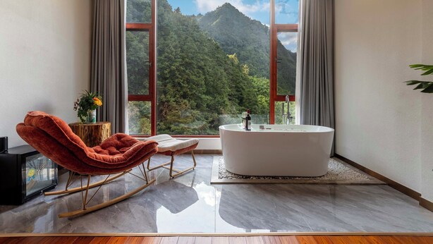 A modern bathroom with a large window, a white bathtub, a red rocking chair, and a small table with a vase. - Huagu Jianzhu Guesthouse Zhangjiajie (Zhangjiajie)