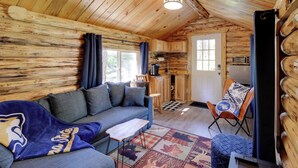 L’intérieur d’un chalet accueillant, avec un canapé, une petite table, un tapis à motifs, un fauteuil et un coin cuisine équipé d’un réfrigérateur et d’un grille-pain.