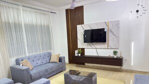 Un salon moderne avec un canapé gris, des fauteuils assortis, une table basse en verre et un téléviseur fixé sur un mur en marbre.