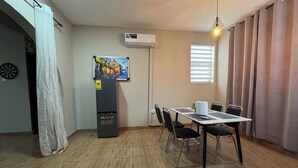 Un espace repas comprenant une table, des chaises, un climatiseur, un réfrigérateur, un tableau accroché au mur et une fenêtre avec des stores.
