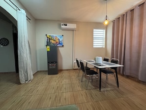 Un comedor con una mesa, sillas, aire acondicionado, refrigerador, un cuadro en la pared y una ventana con persianas.