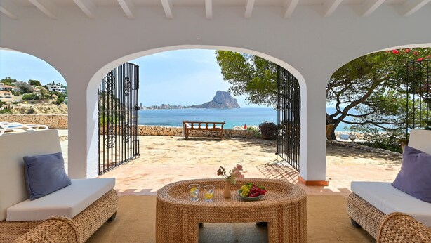 A beachside patio with wicker furniture, a round table with drinks and fruit, and a view of the sea and a mountain. - Casa Timeris - Calpe, Costa Blanca (Calpe)