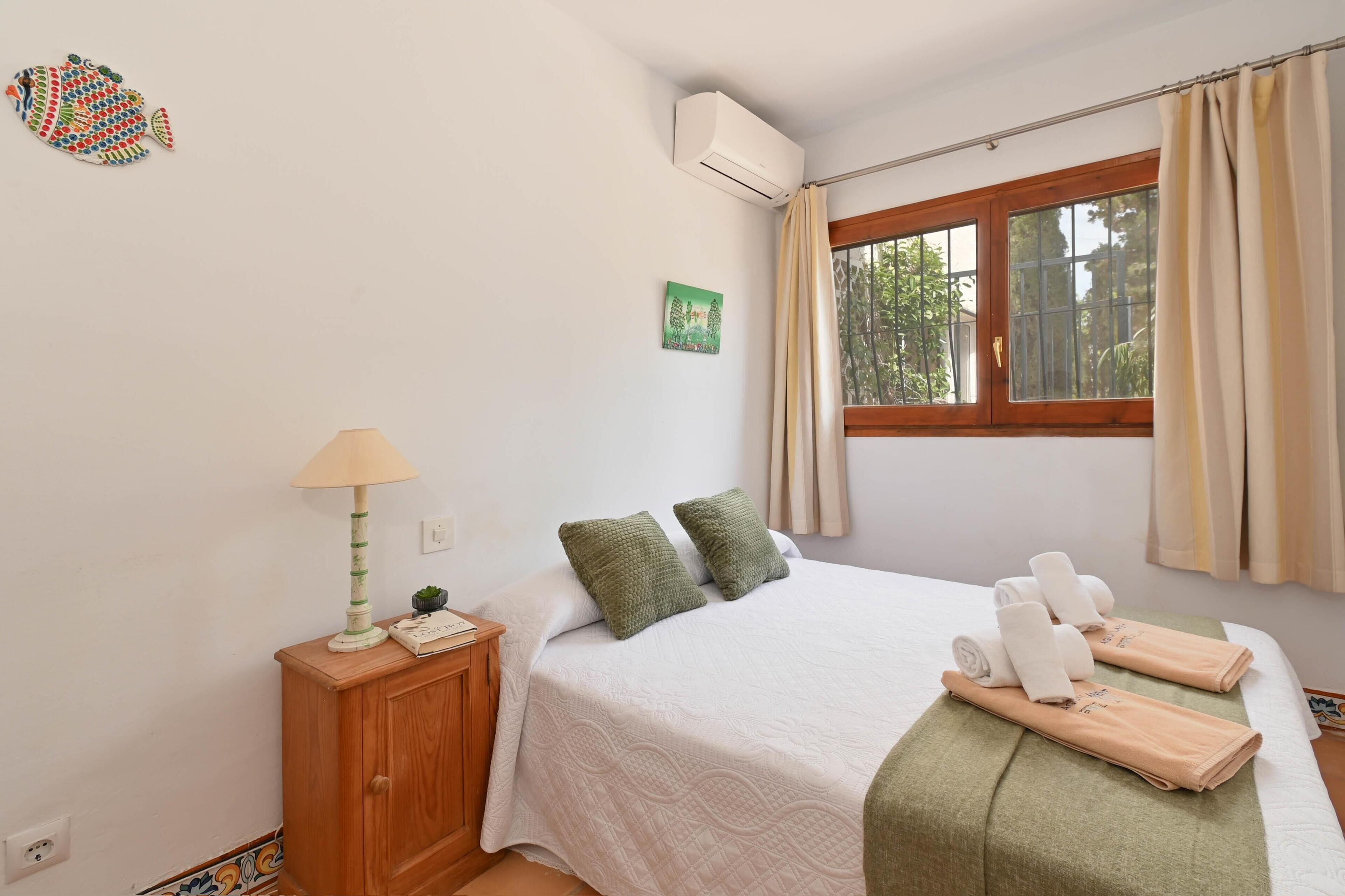 A neatly arranged bedroom with a bed, pillows, a bedside table with a lamp, a window with curtains, and a wall-mounted air conditioner.