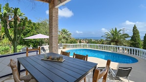 Un espace aménagé au bord de la piscine, avec une table et des chaises, entouré d’une balustrade blanche et d’une végétation luxuriante.
