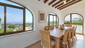 Un coin repas avec une table en bois et des chaises en osier, de grandes fenêtres en arc de cercle et une vue sur le paysage.