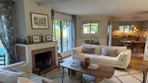 Un salón amplio con chimenea, un sofá, una mesa de centro y un área de comedor visible a través de puertas de cristal.