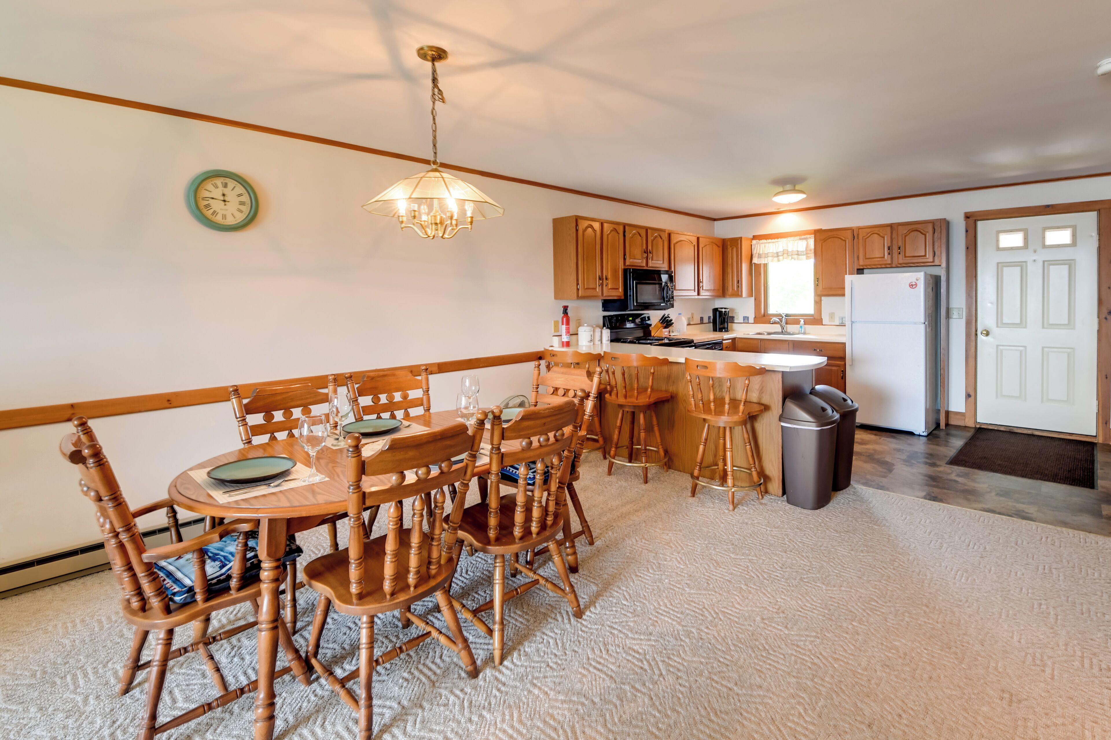 A dining area with wooden chairs and a table, a kitchen with wooden cabinets and appliances, and a clock on the wall.