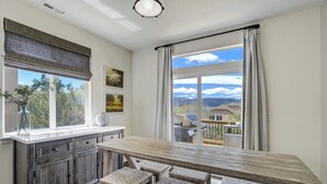Cozinha com mesa de madeira e banquetas, janelas amplas com cortinas e vista para o exterior.