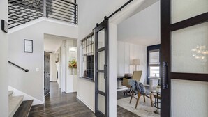 Um corredor espaçoso com piso de madeira escura, escada e uma grande porta de vidro deslizante que dá para uma sala de estar com escrivaninha e cadeiras.