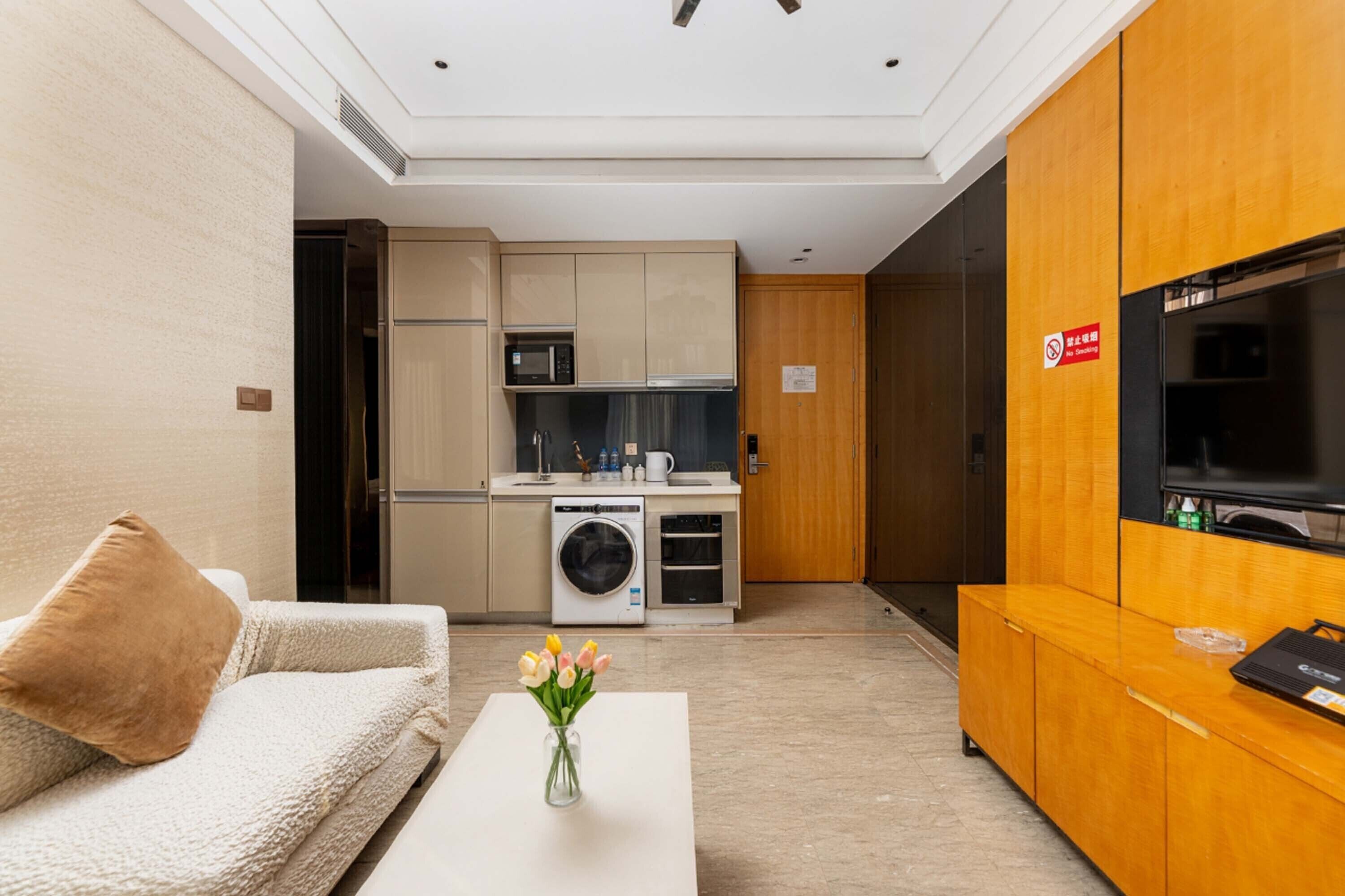 Ein modernes Hotelzimmer mit einer Couch, einem Couchtisch mit Blumenvase, einer Küchenzeile inklusive Mikrowelle, Backofen und Waschmaschine sowie einem Fernseher auf einer Holzkommode.