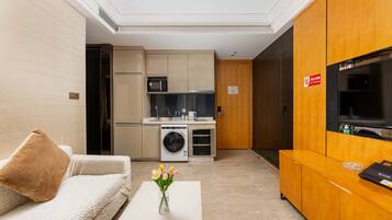 A modern hotel room with a sofa, a coffee table with a vase of flowers, a kitchen area with a microwave, oven, and washing machine, and a television on a wooden cabinet.