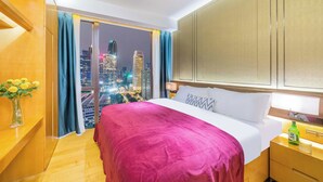 Ein Hotelzimmer mit einem großen Bett, einem Nachttisch, einer Blumenvase und Blick auf eine nächtliche Stadtlandschaft.