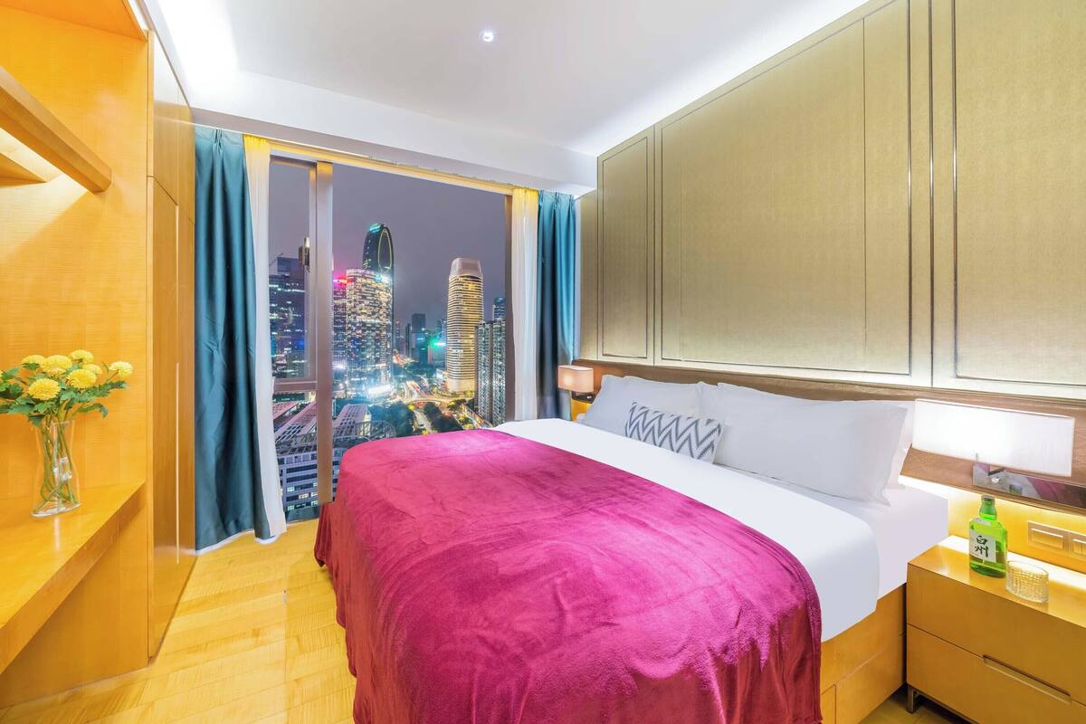 A hotel room with a large bed, a nightstand, a vase of flowers, and a view of a cityscape at night.