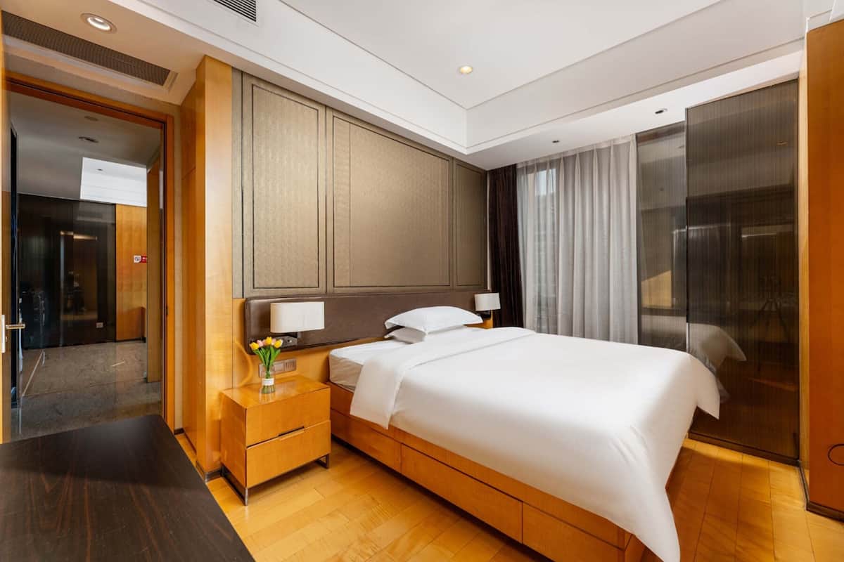 A modern hotel room with a large bed, wooden nightstand, and a glass partition wall.