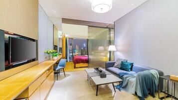A modern hotel room with a flat-screen TV, a sofa, a coffee table, a desk, and a view of the cityscape through the window.