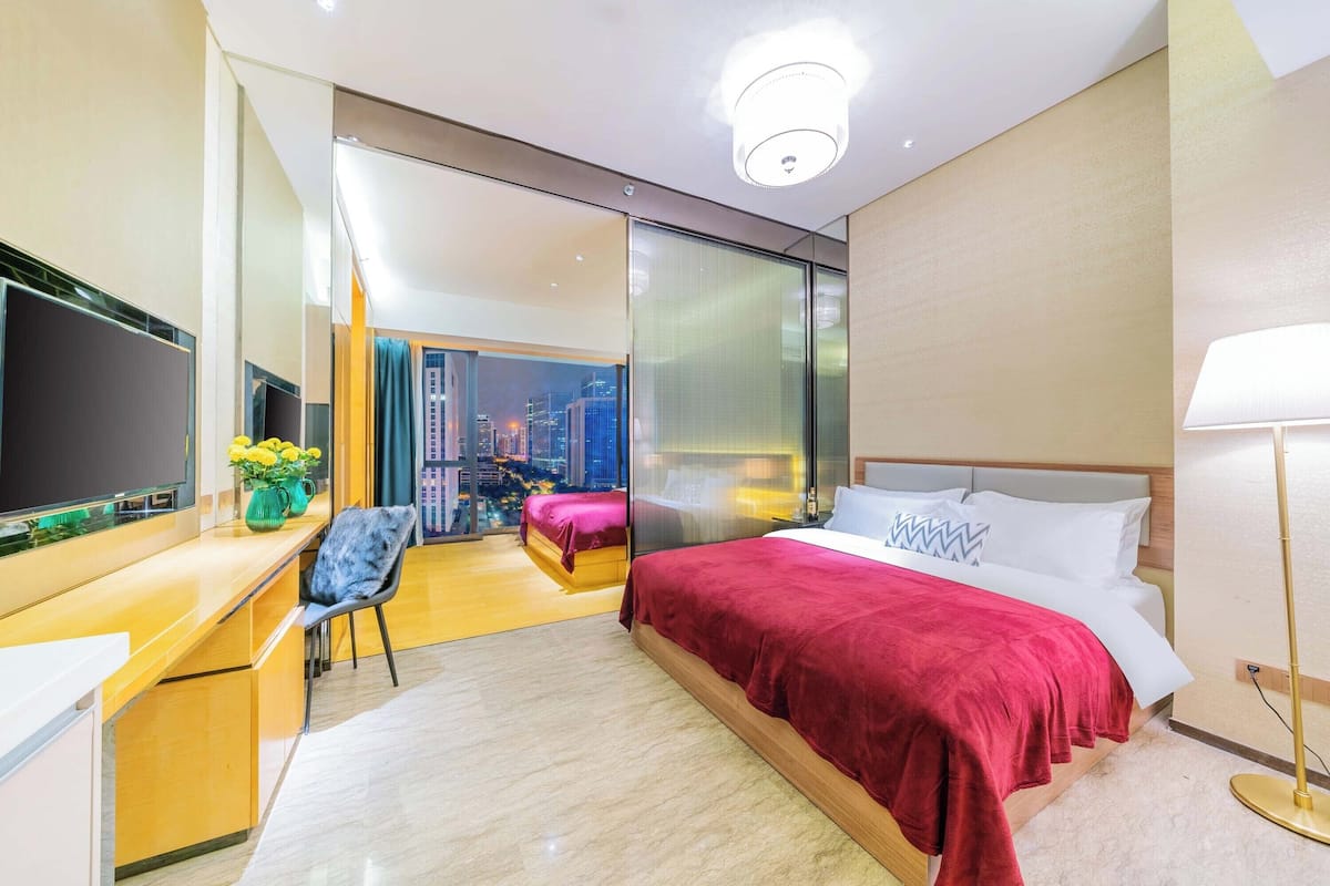 A modern hotel room with a large bed, a desk with a computer, a chair, and a view of a cityscape through a glass door.