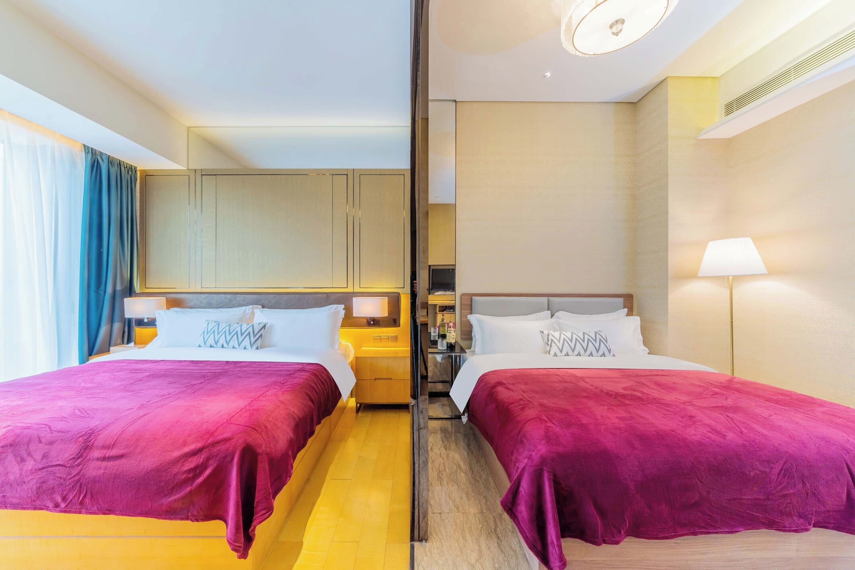 A hotel room with two beds, a nightstand, a lamp, and a view of the city.