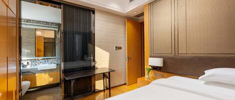 Una habitación de hotel con cama, escritorio, espejo, baño con lavamanos e inodoro, y cabecera de madera.