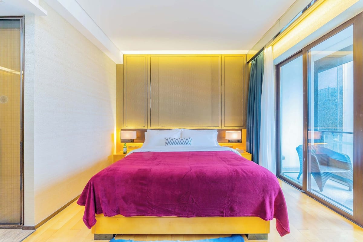 A modern hotel room with a large bed, a sliding glass door to a balcony, and a view of the cityscape.