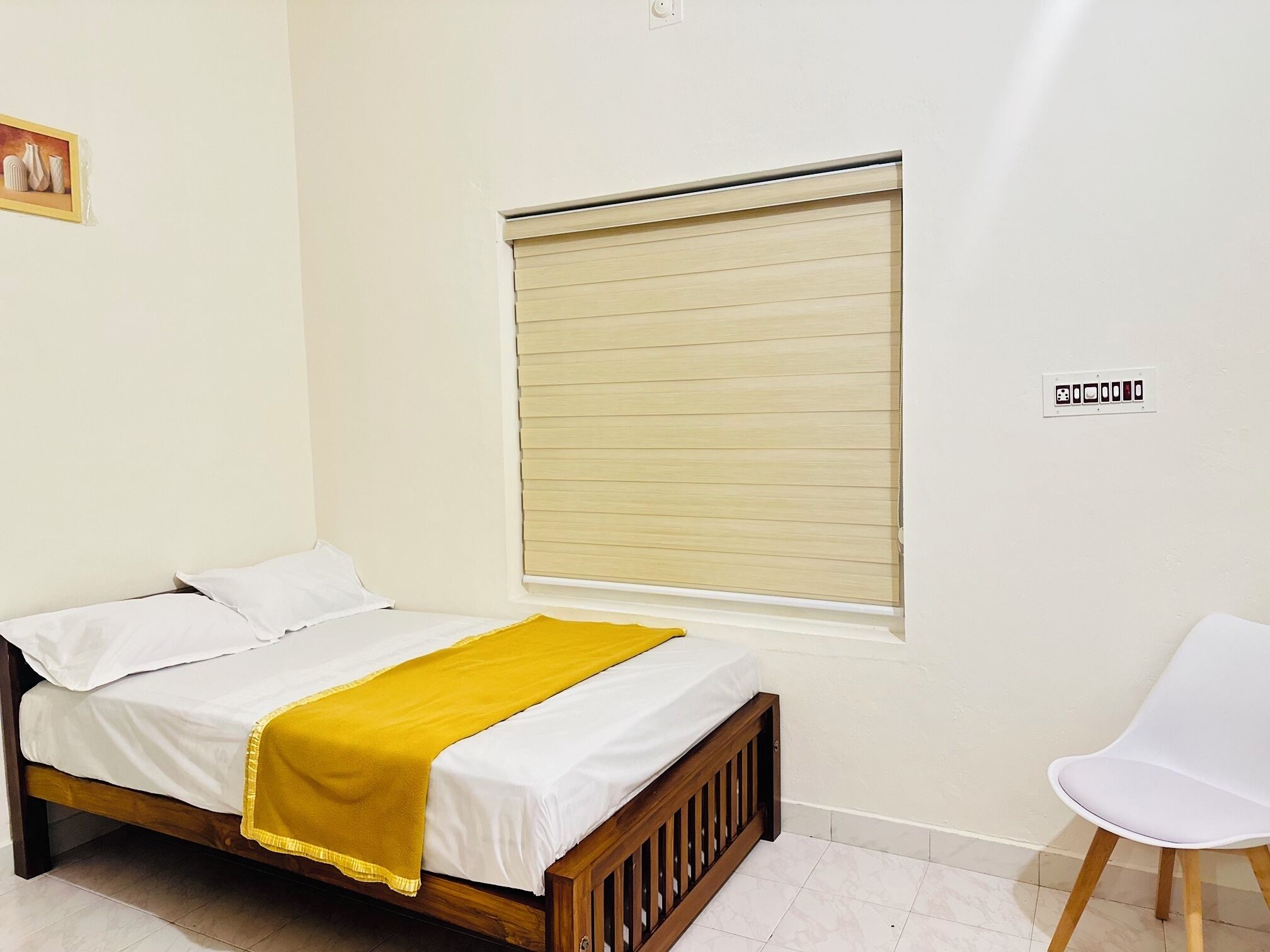 Una cama individual con una manta amarilla, una silla blanca y una ventana con persianas.