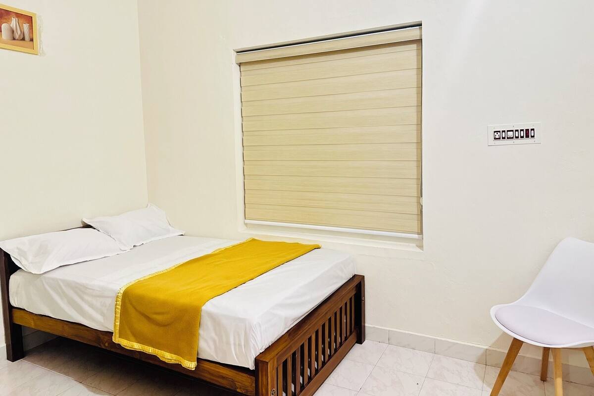 A single bed with a yellow blanket, a white chair, and a window with blinds.