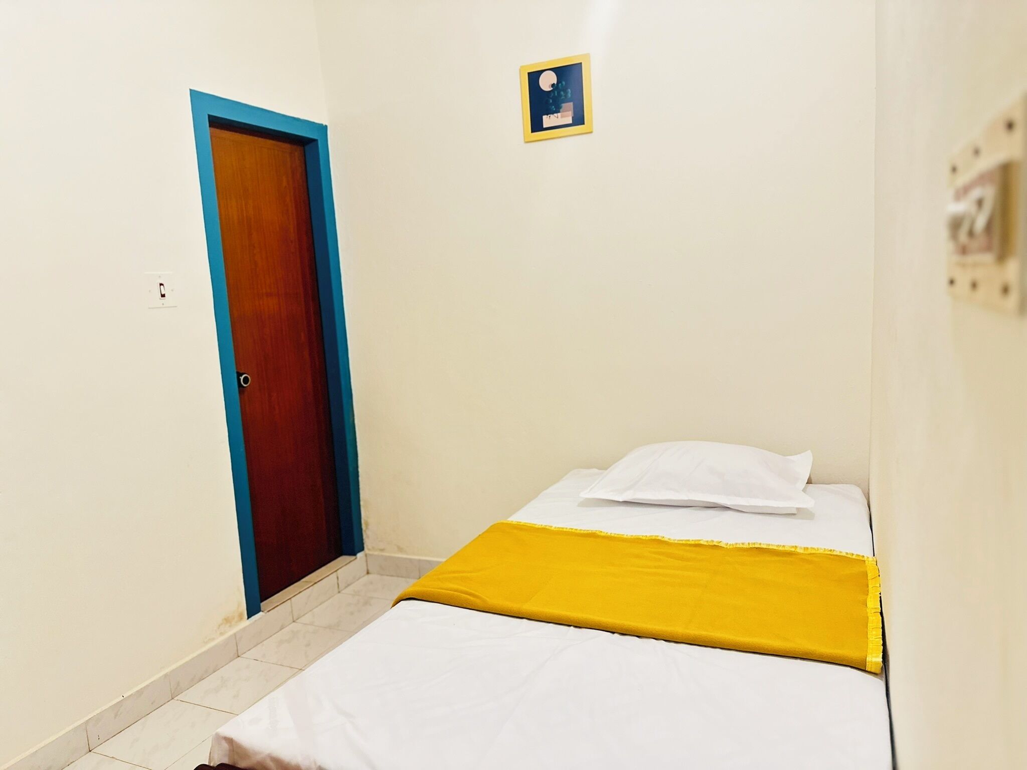 Una habitación pequeña con una cama individual, una puerta de madera, una cama blanca con una manta amarilla y un cuadro pequeño en la pared.