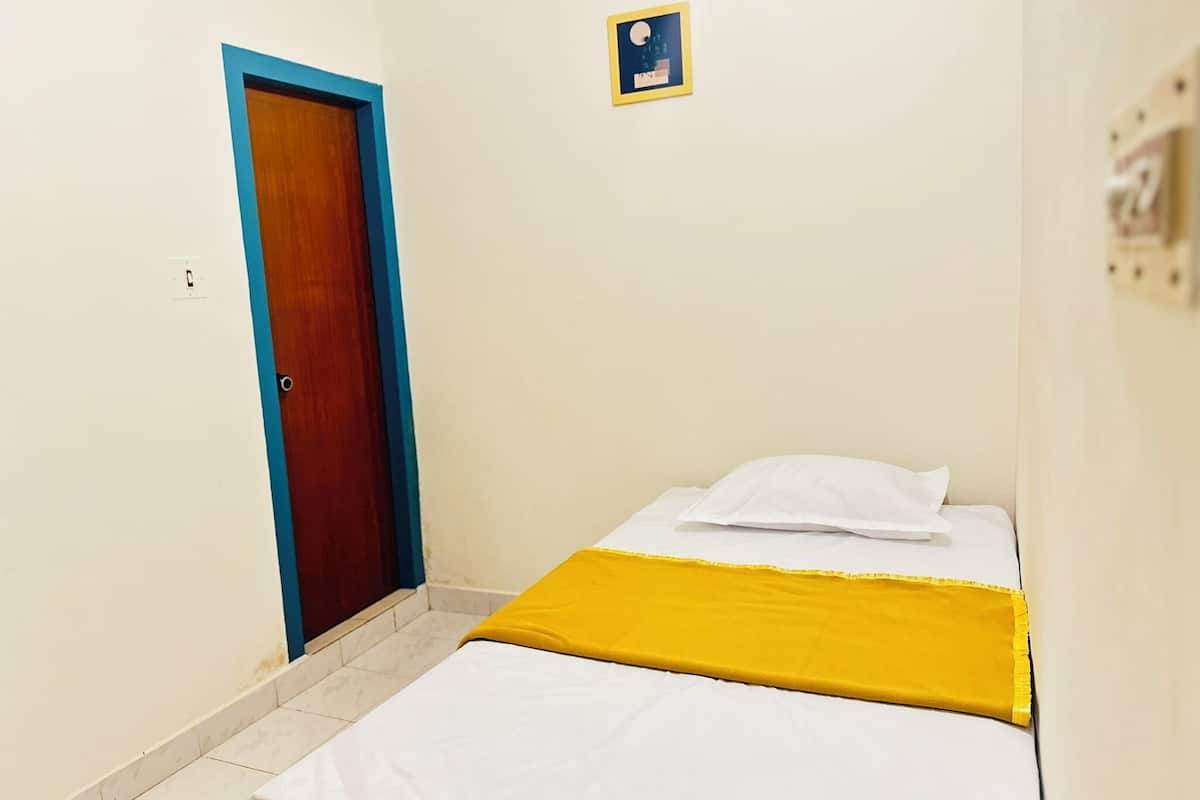 A small, single-bed room with a wooden door, a white bed with a yellow blanket, and a small framed picture on the wall.