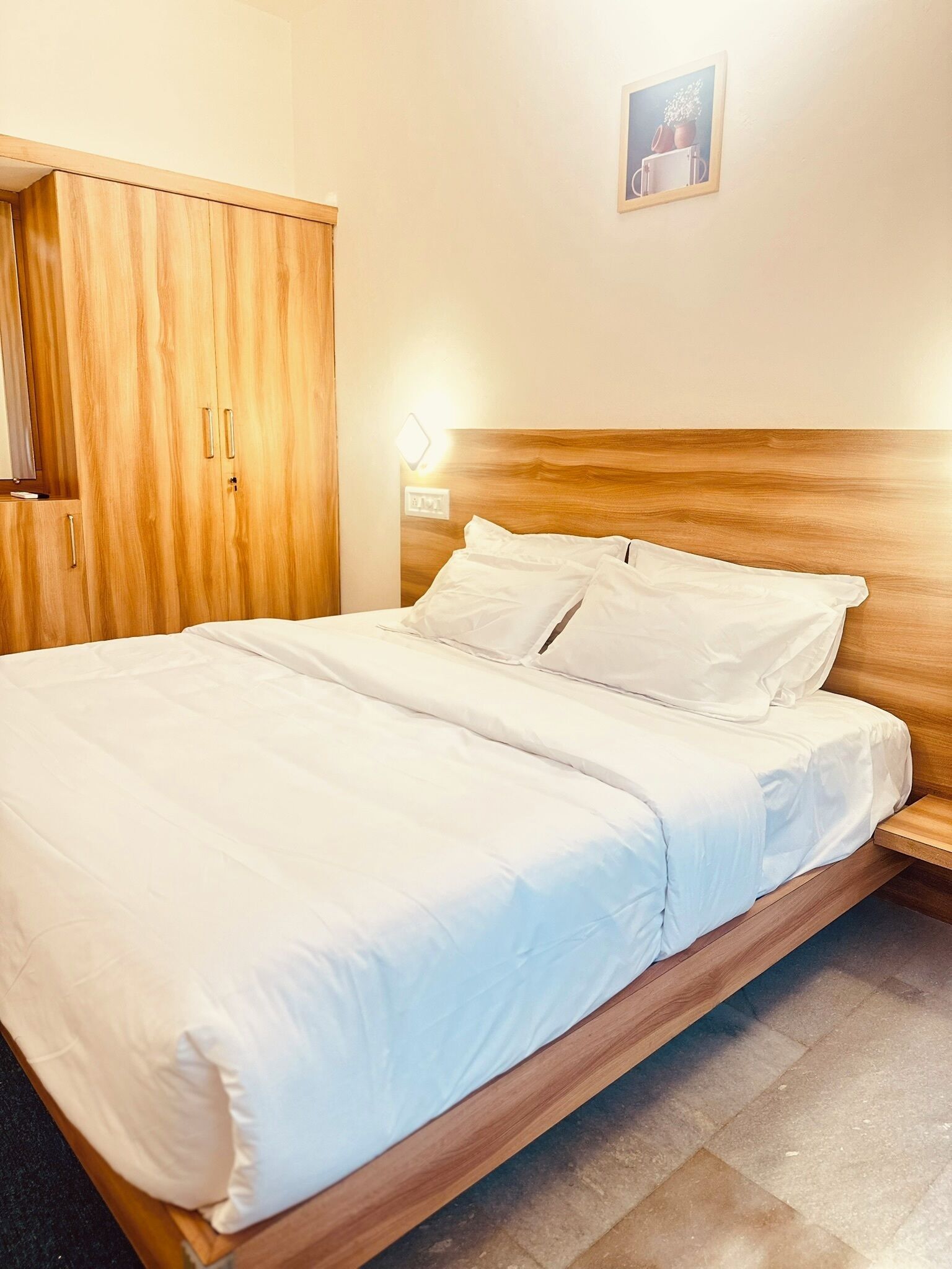 Una cama bien tendida con sábanas blancas en una habitación con muebles de madera y una lámpara pequeña fijada a la pared.