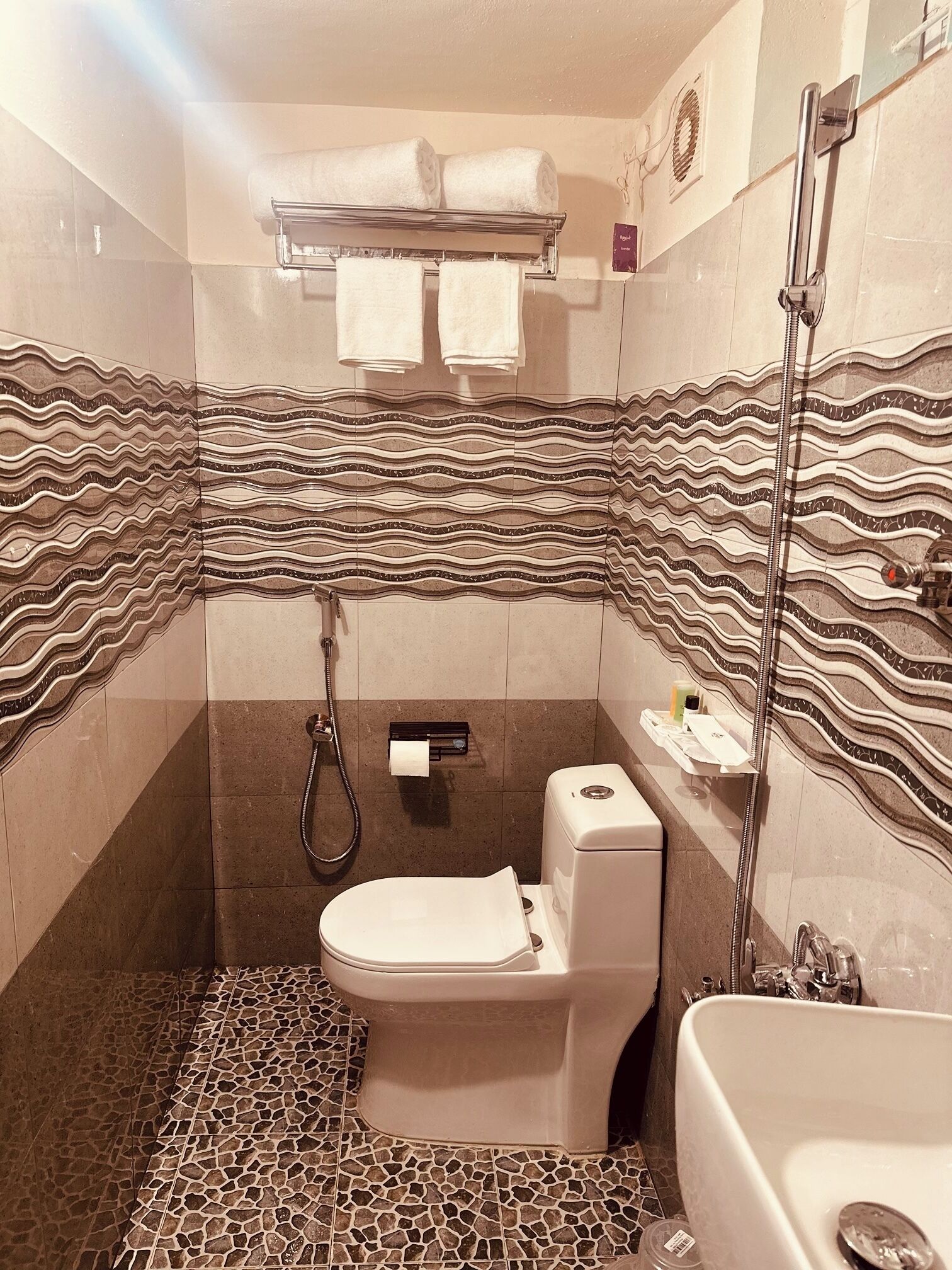 Et badeværelse med flisegulv, toilet, brusebad og et håndklædestativ med håndklæder.