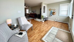 A living room with a sofa, a coffee table, and a lamp. There is a dining area with a table and chairs. The room has wooden flooring and a large window. - 5BR Sandy Pond MAIN LAKE HOUSE | Dock | Fire Pit | Pets (Sandy Creek)