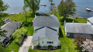 A large house with a steep roof, surrounded by a well-maintained lawn and trees, overlooking a calm lake with boats. - 5BR Sandy Pond MAIN LAKE HOUSE | Dock | Fire Pit | Pets (Sandy Creek)