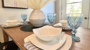 Una mesa de comedor dispuesta con platos blancos, cuencos y cristalería azul, un jarrón con plumas blancas y un cuadro en la pared.