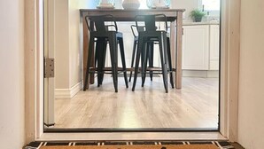 Una cocina moderna con comedor, que incluye una mesa de madera y sillas negras, y un felpudo con la inscripción 'gay! you're here!'.