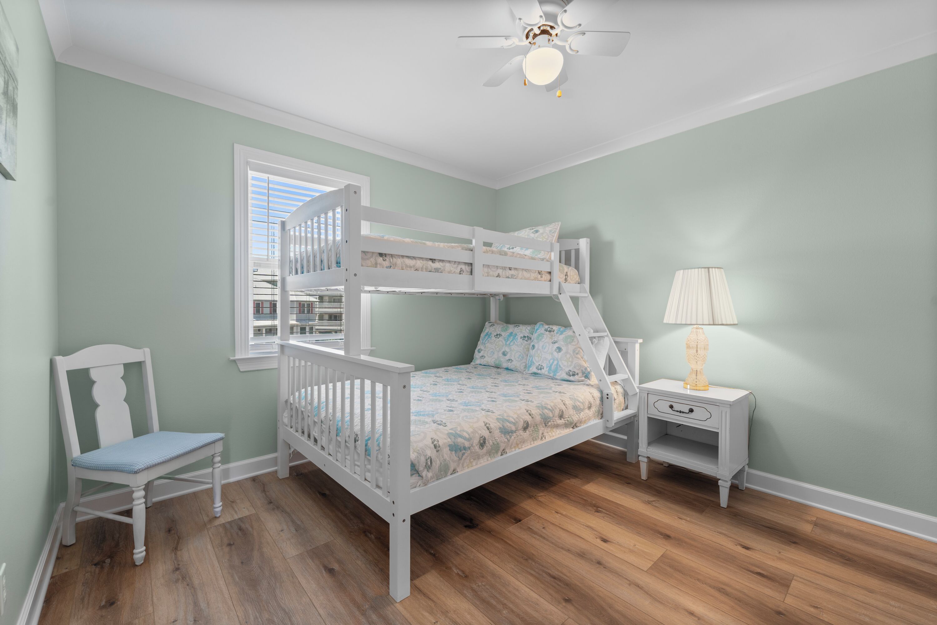 A bunk bed room with a ceiling fan, a nightstand with a lamp, a chair, and a window with blinds.