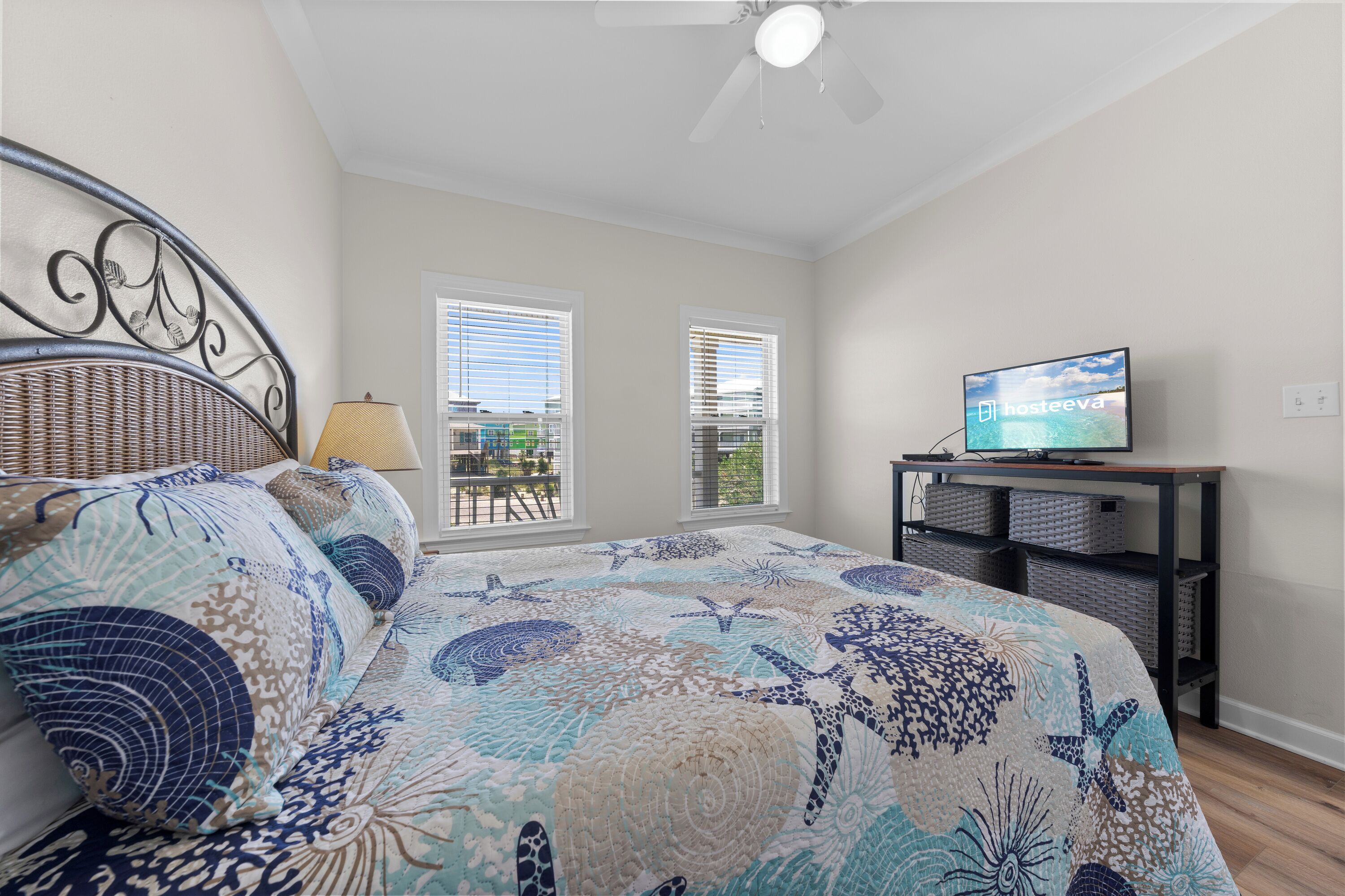 A bedroom with a bed, a television on a stand, two windows, and a ceiling fan.