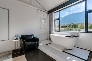 A modern bathroom with a bathtub, a black leather chair, a small table with water bottles, and a large window offering a mountain view.