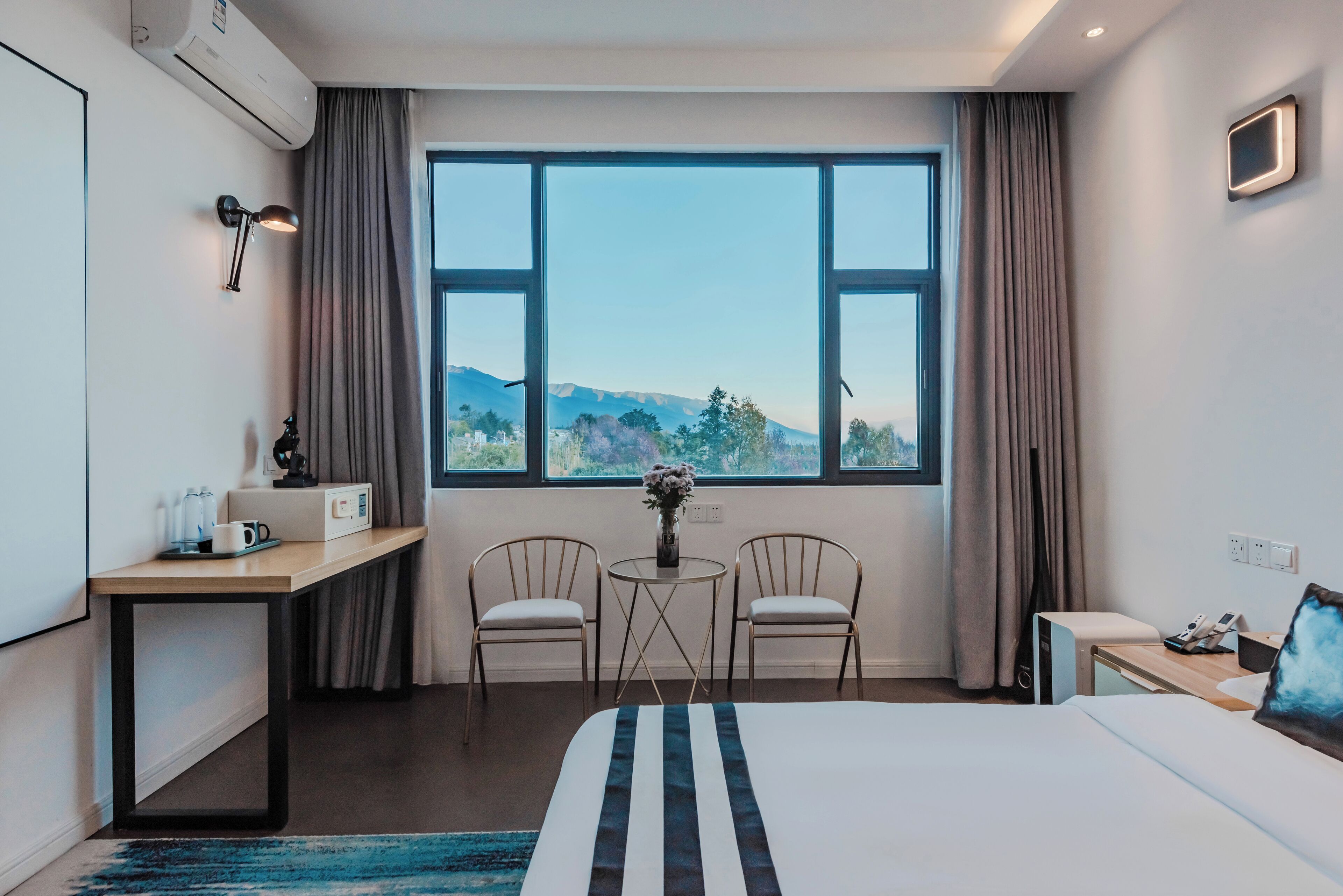 A hotel room with a bed, a desk, two chairs, a small table with a vase, and a large window with a mountain view.
